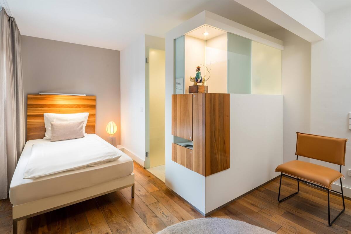 A modern hotel room with a wooden floor, a bed with white linens, a wooden cabinet, and a brown leather chair.