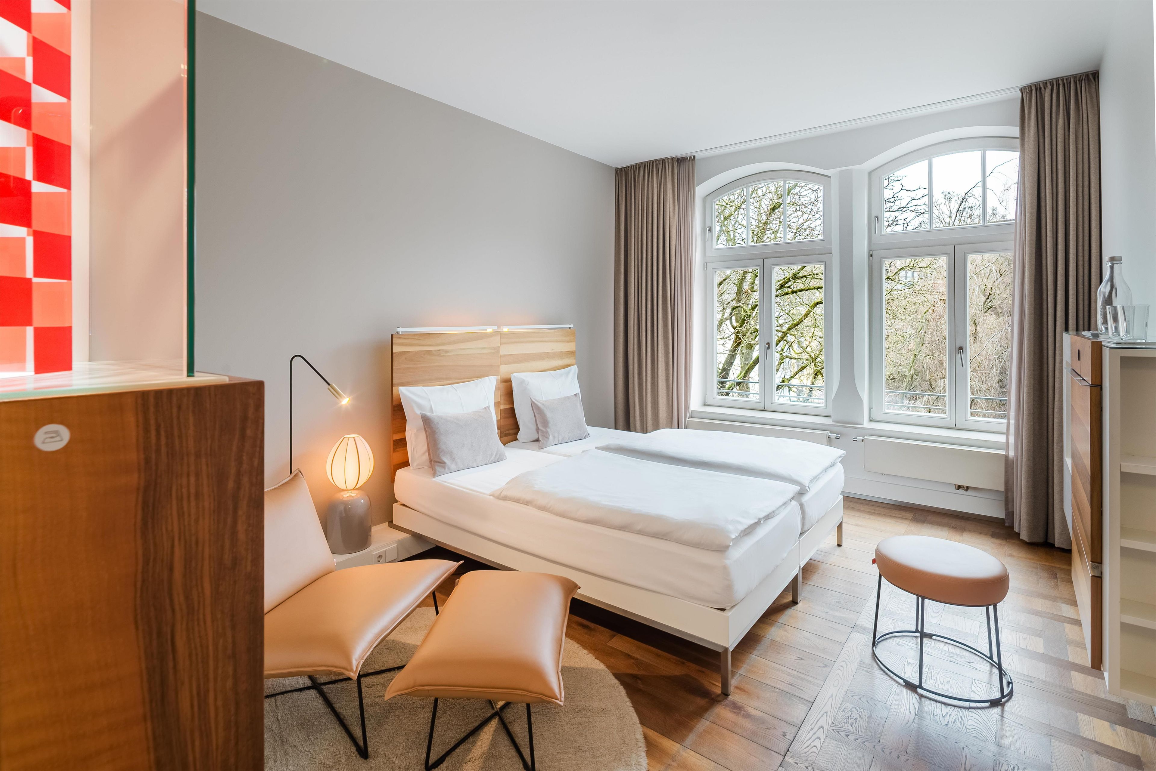 A modern bedroom with a large bed, bedside tables, a desk, and a view of trees through the window.