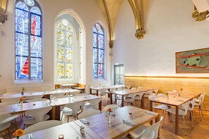 Een eetruimte met meerdere tafels en stoelen, grote glas-in-loodramen en een hoge plafond met decoratieve details.
