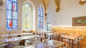 A dining area with tables and chairs, large stained glass windows, and a painting on the wall. - Hopper Hotel St. Josef, WorldHotels Crafted (Cologne)