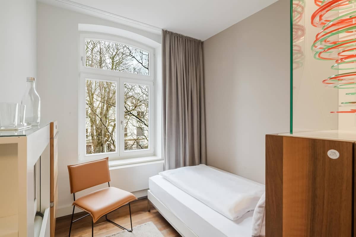 A bedroom with a bed, a chair, a desk with a lamp, a window with curtains, and a wall art piece.