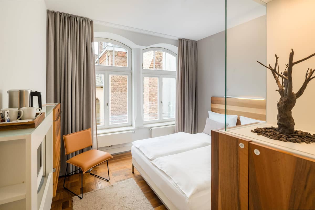 A hotel room with a bed, a desk with a kettle, a chair, and a large window with curtains.