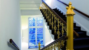 Una escalera con escalones negros y balaustres dorados, que conduce a un rellano con un ventanal azul.