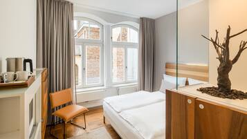 A hotel room with a bed, a desk with a kettle, a chair, and a view of a brick building through the window.