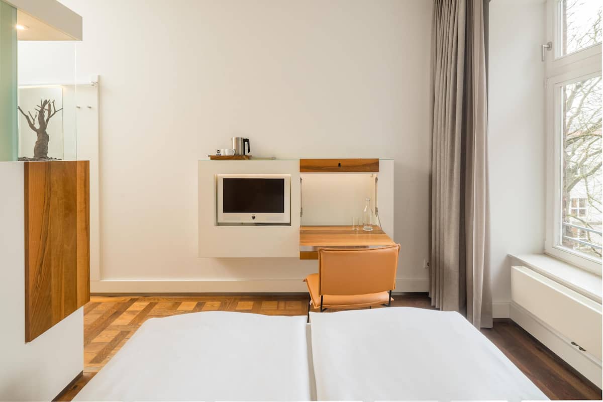 A hotel room with a bed, a TV, a desk with a lamp, a chair, and a window with curtains.