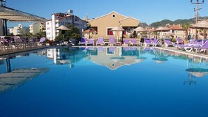 A swimming pool with purple lounge chairs and umbrellas, surrounded by buildings with a Mediterranean architectural style. - Reis Maris Hotel (Marmaris)