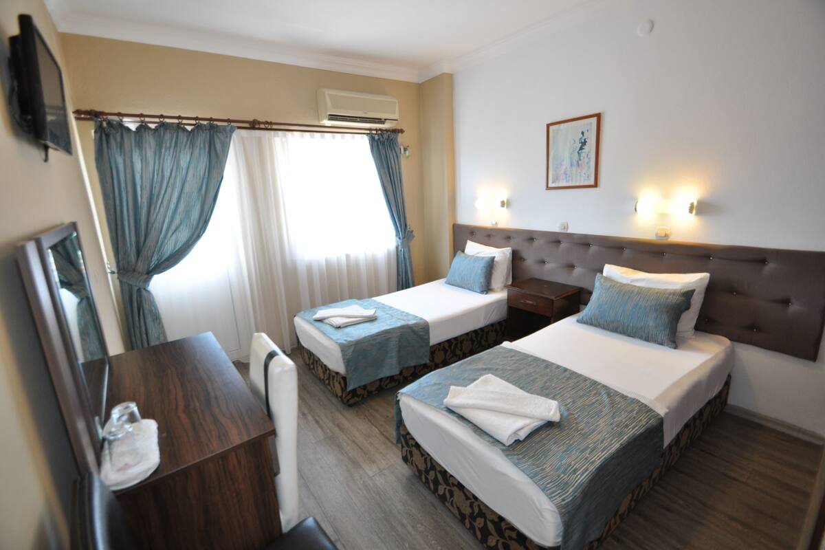 A hotel room with two single beds, a desk, a chair, a mirror, and a painting on the wall.