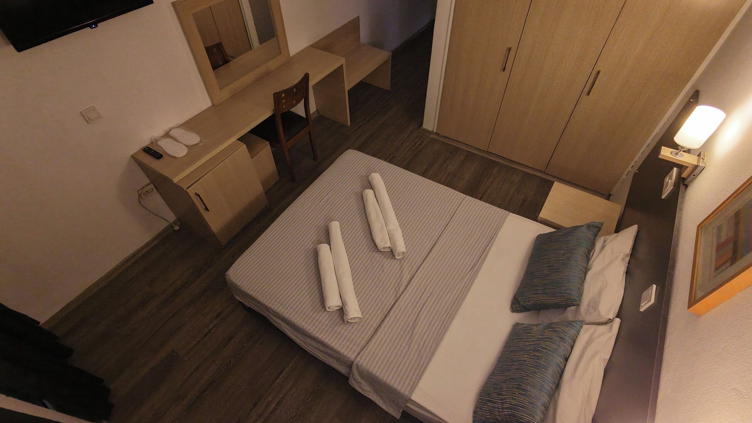 A hotel room with a bed, two rolled towels, a desk, a chair, and a wardrobe.
