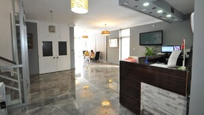 A reception area with a dark wooden desk, a flat-screen TV, and a potted plant. There are two pendant lights hanging from the ceiling, and a staircase is visible to the left. - Reis Maris Hotel (Marmaris)