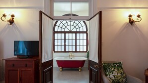 A bathroom with a freestanding bathtub, a television on a wooden cabinet, and a chair with a floral pillow. - Esperanza Guest House (Cape Town)