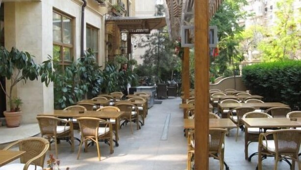 An outdoor cafe area with wicker tables and chairs, potted plants, and a covered walkway. - Hotel Irisa (Bucharest)