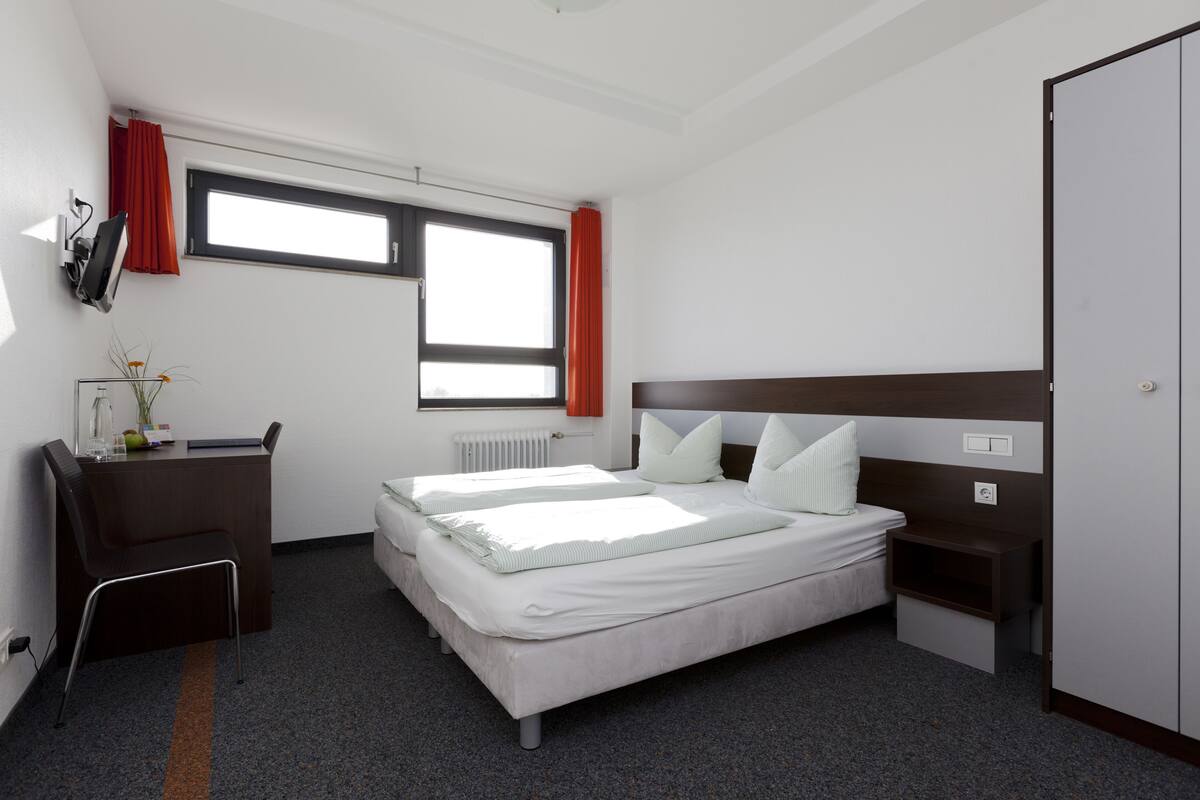 Habitación de hotel con cama, escritorio, silla, armario y ventana con cortinas.