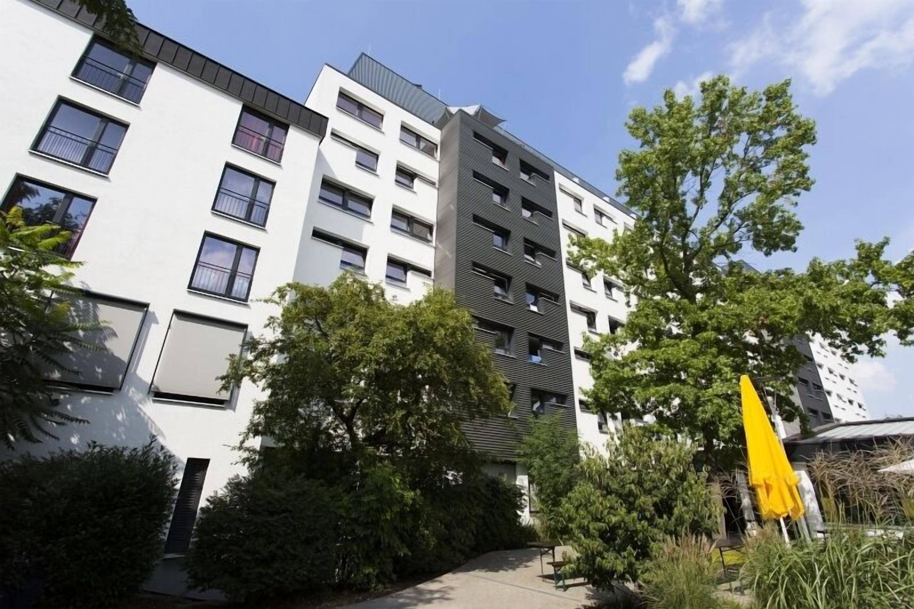 A multi-story residential building with balconies, surrounded by greenery and a yellow umbrella.