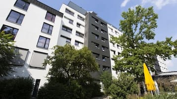 A multi-story residential building with balconies, surrounded by greenery and a yellow umbrella.