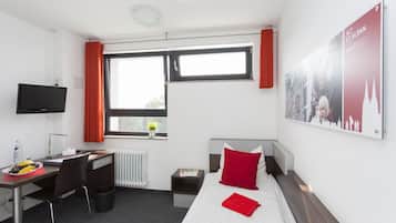 A hotel room with a bed, a desk, a chair, a TV, a window with curtains, a radiator, and a poster on the wall.