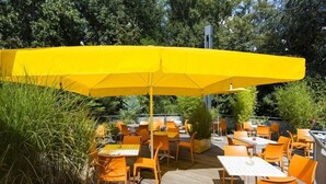 An outdoor patio with orange chairs and tables, a yellow umbrella, and greenery. - DJH City-Hostel Köln-Riehl (Cologne)
