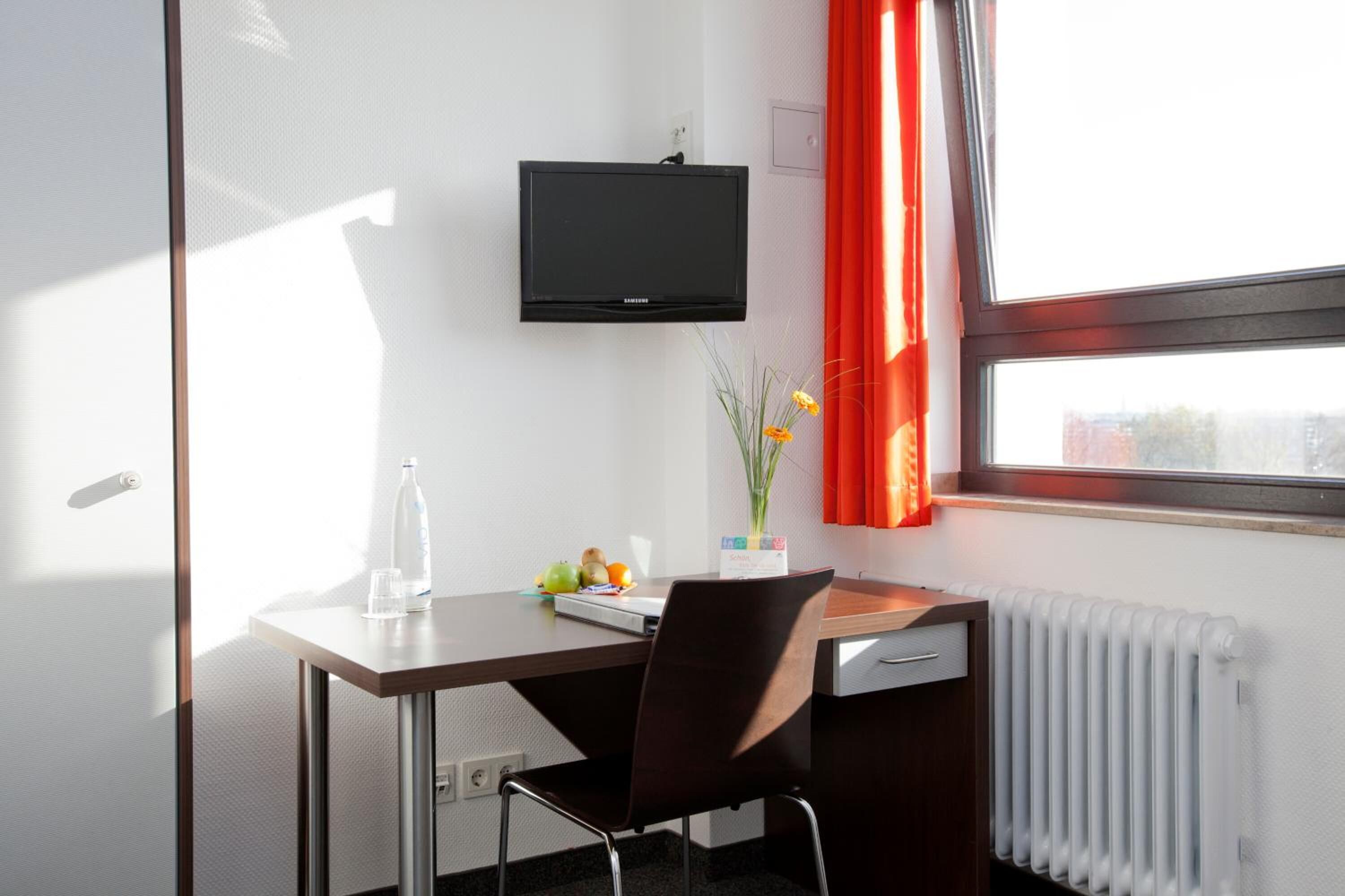 Una habitación de hotel con escritorio, silla, televisor, radiador, ventana con cortinas y un jarrón con flores.