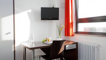 Una habitación de hotel con escritorio, silla, televisor, radiador, ventana con cortinas y un jarrón con flores.