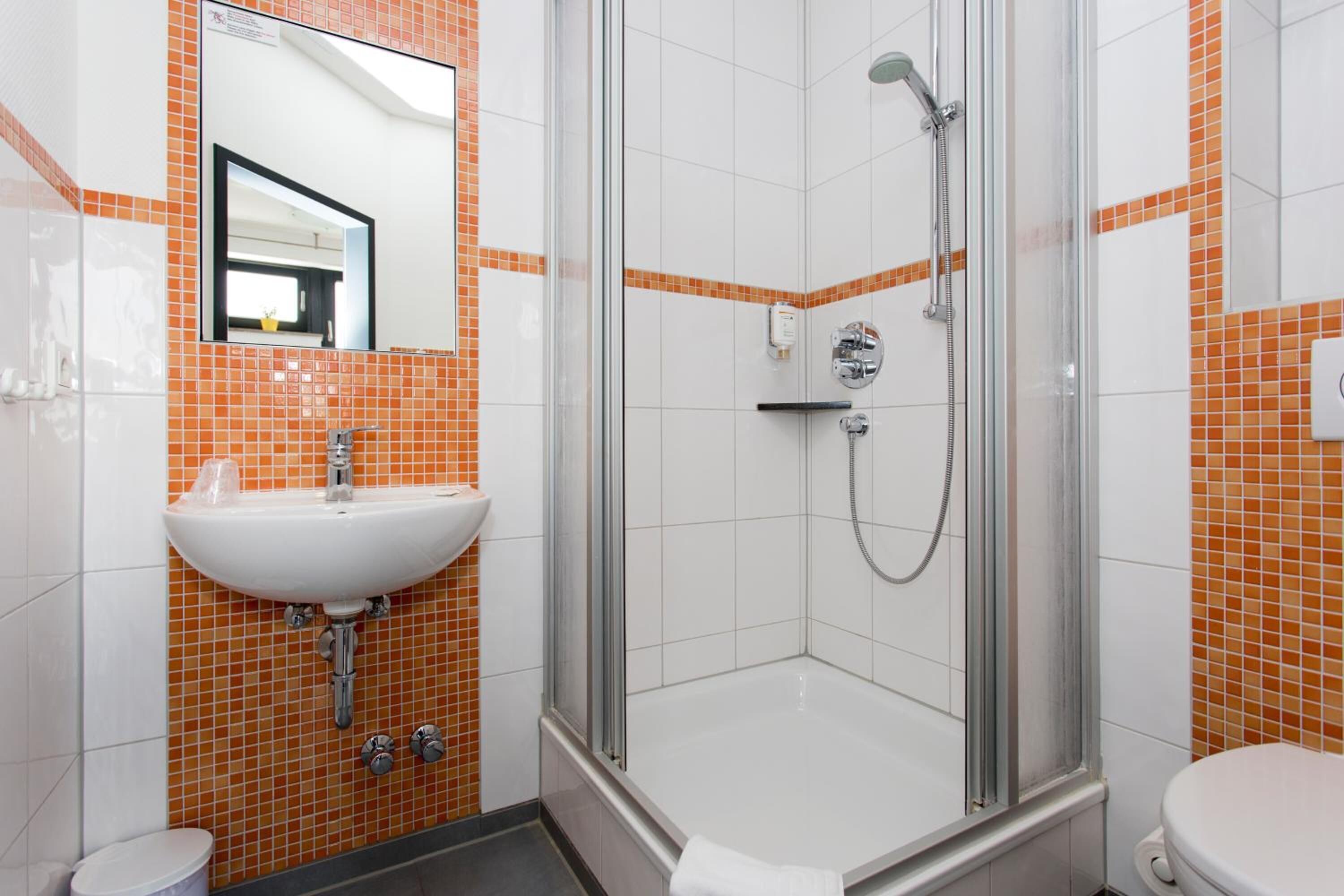 Un baño moderno con ducha envuelta en vidrio, un lavamanos blanco y paredes con azulejos naranja.