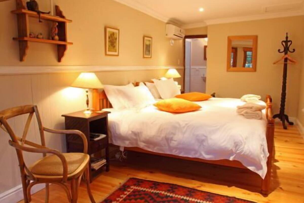 A neatly arranged bedroom with a wooden bed, white bedding, a wooden chair, a bedside table with a lamp, and a small shelf with decorative items.