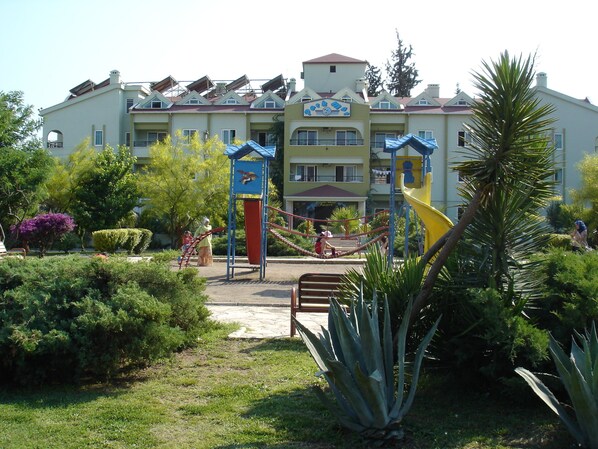 A multi-story building with a playground in front, surrounded by greenery and a palm tree. - Park Mar Apart (Marmaris)