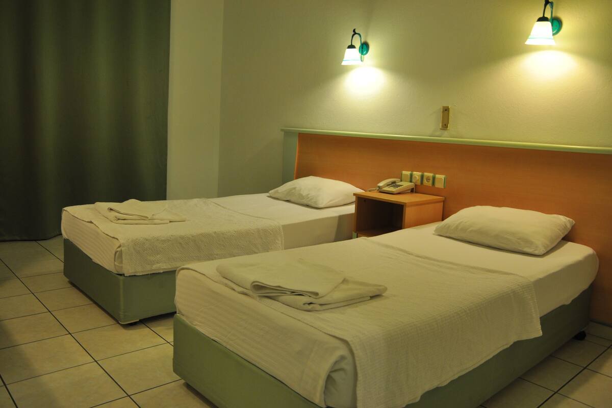 Two twin beds with white linens, a bedside table with a lamp, and a curtain on the left.