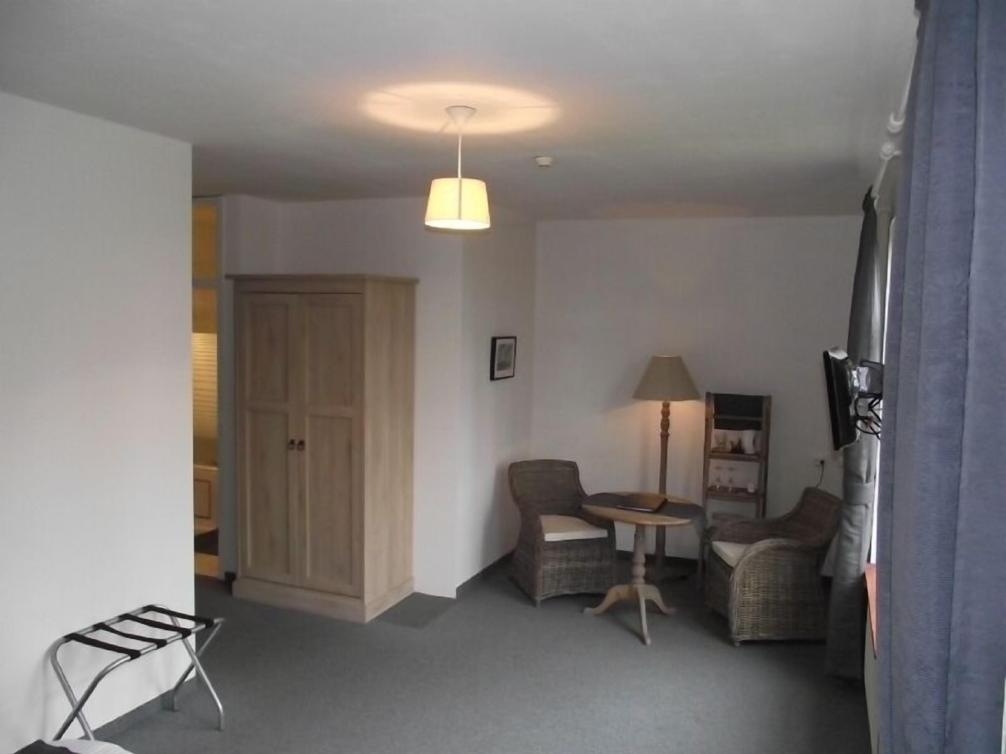 A small, cozy room with a wooden wardrobe, two wicker chairs, a small table, and a hanging light fixture.