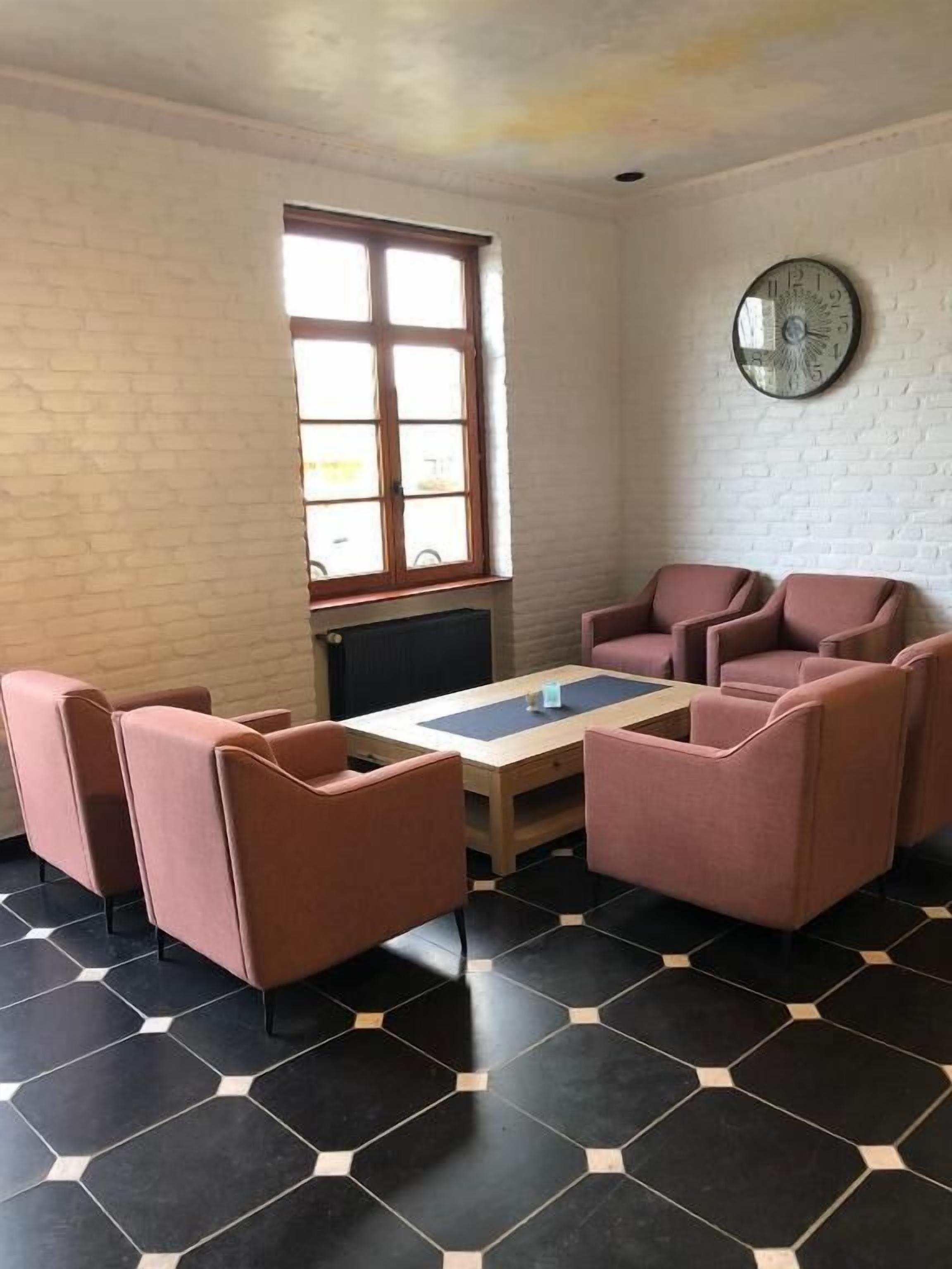 Een kamer met een bakstenen muur, een raam, een ronde wandklok en een zithoek met een tafel en stoelen.