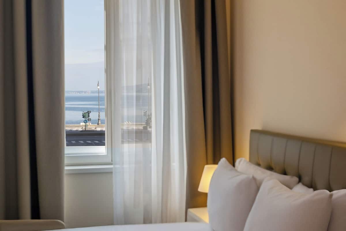 A hotel room with a bed, a window with curtains, and a view of the sea.