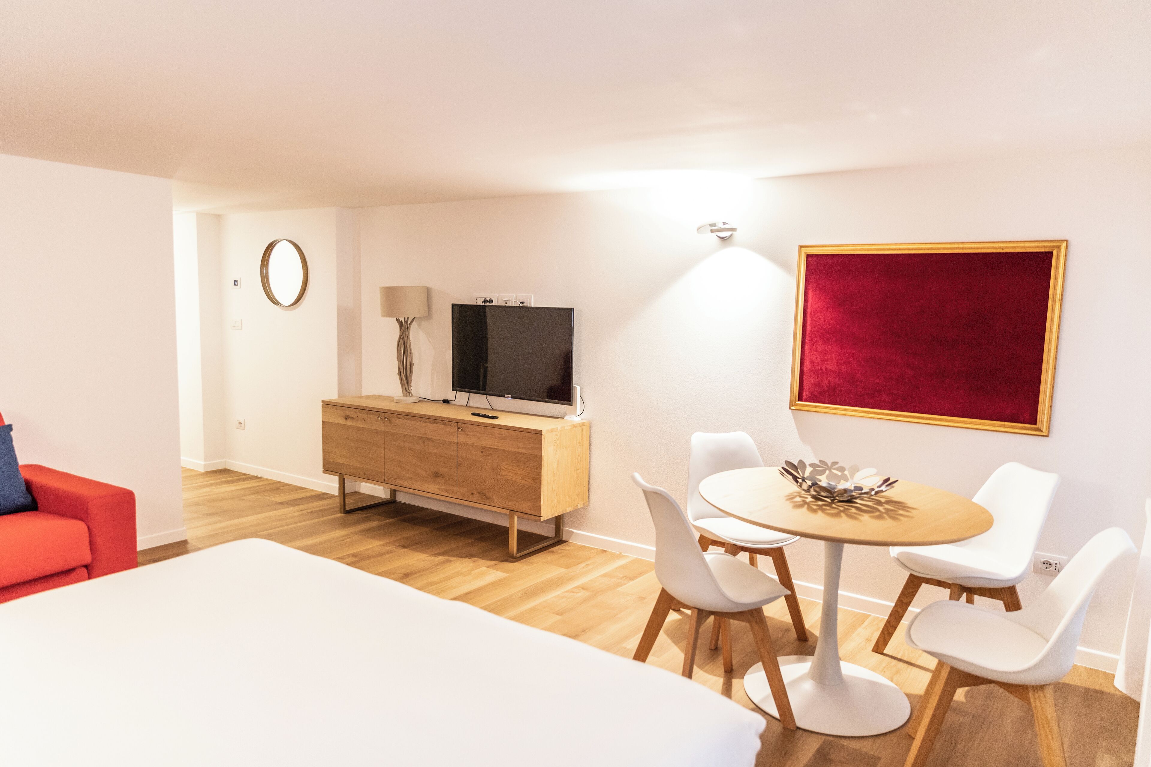 Quarto de hotel moderno com penteadeira de madeira, televisão de tela plana, mesa de jantar redonda com cadeiras brancas, parede com detalhe vermelho e pintura aveludada em vermelho.