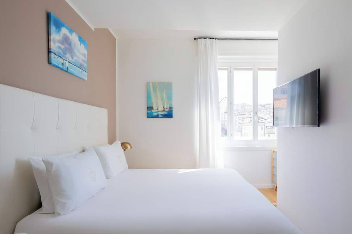 A hotel room with a large bed, a flat-screen TV, two paintings on the wall, and a window with curtains.