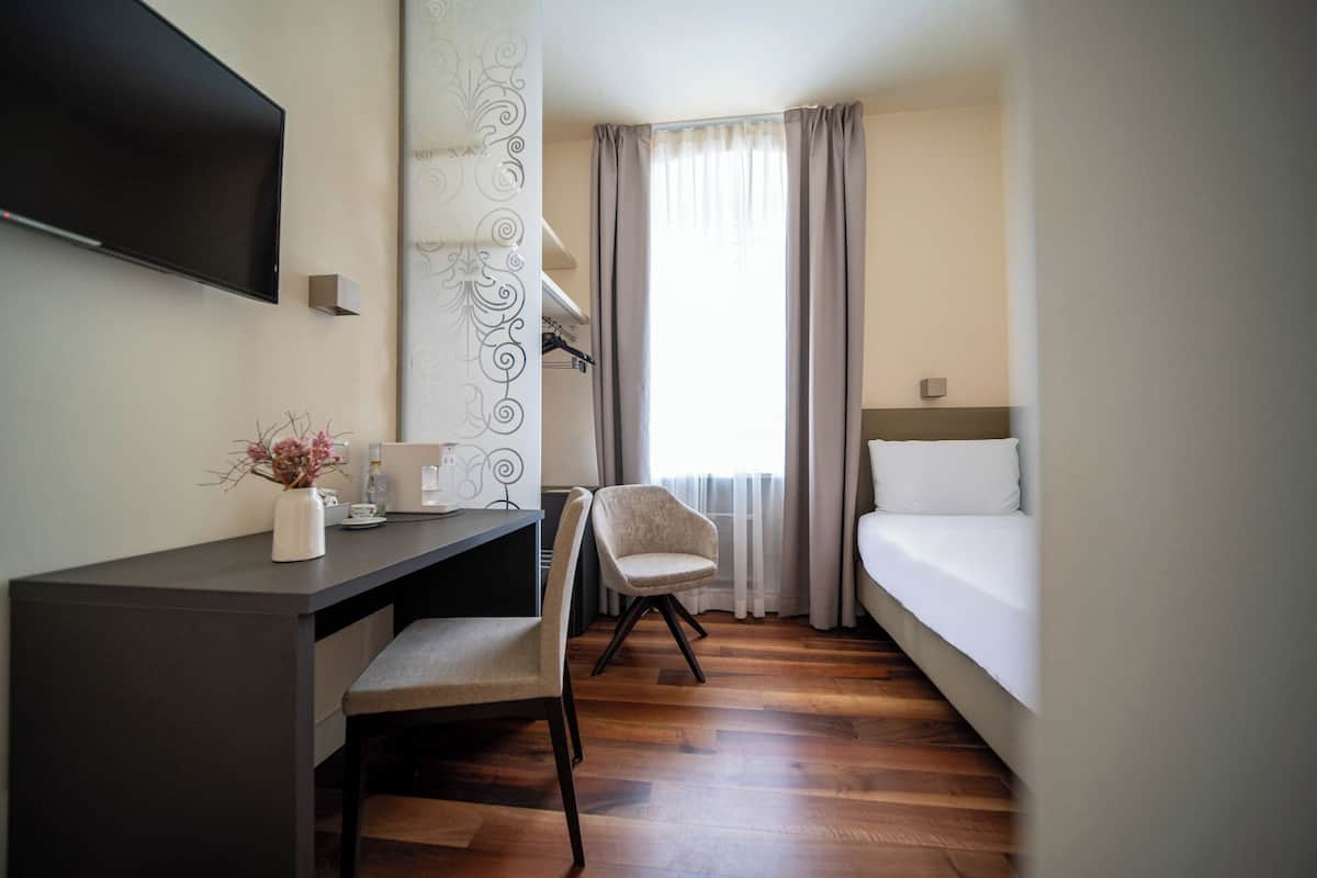 A hotel room with a bed, a desk with a chair, a TV mounted on the wall, and a window with curtains.
