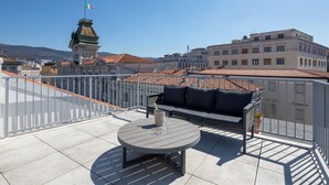 A rooftop terrace with a round table and a sofa, a flag, and buildings in the background. - Hotel Vis a Vis (Trieste)
