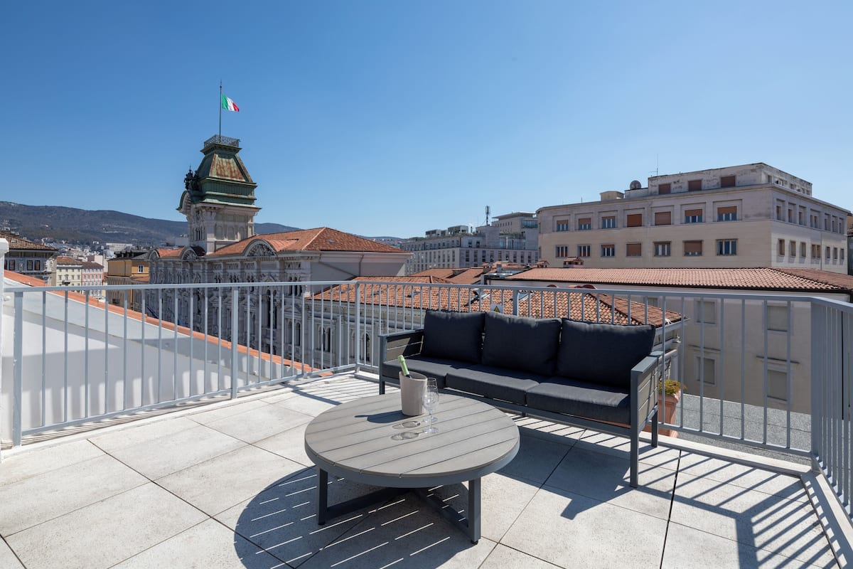 A rooftop terrace with a round table and a sofa, a flag, and buildings in the background.