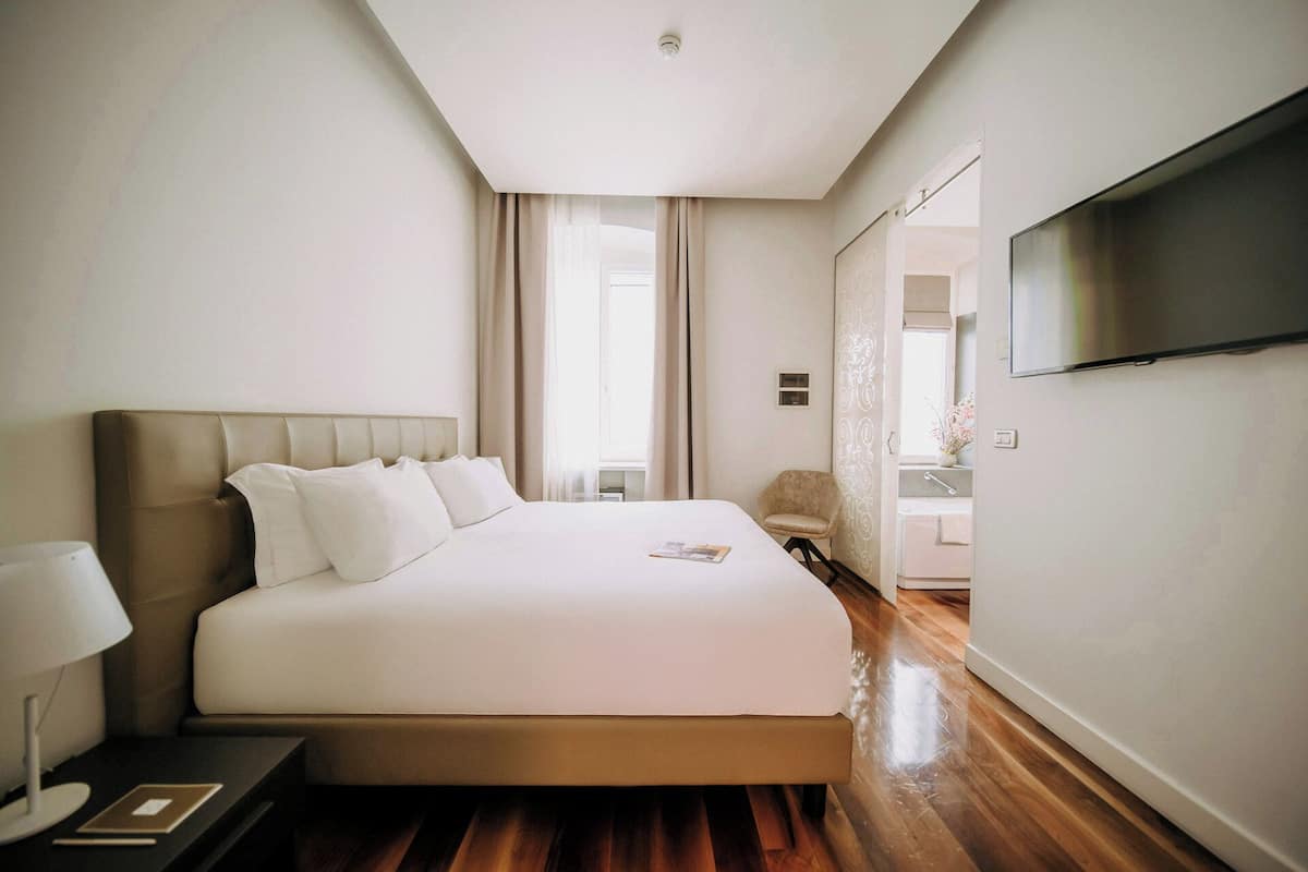 A modern hotel room with a large bed, a flat-screen TV, a bedside table with a lamp, and a view of the bathroom.