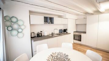 A modern kitchen with white cabinets, a stainless steel sink, a stove, and a coffee maker. There is a dining table with chairs in the foreground.