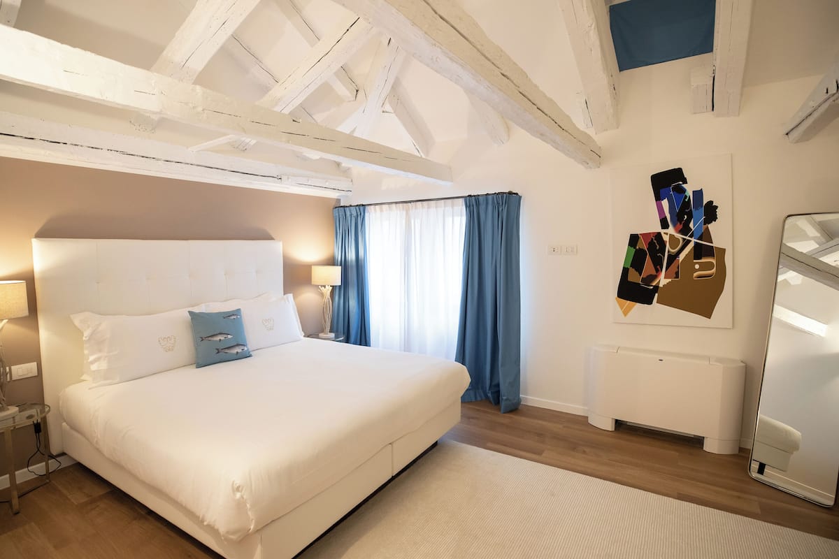 A bedroom with a large bed, a nightstand, a lamp, a radiator, and a wall with a geometric artwork.