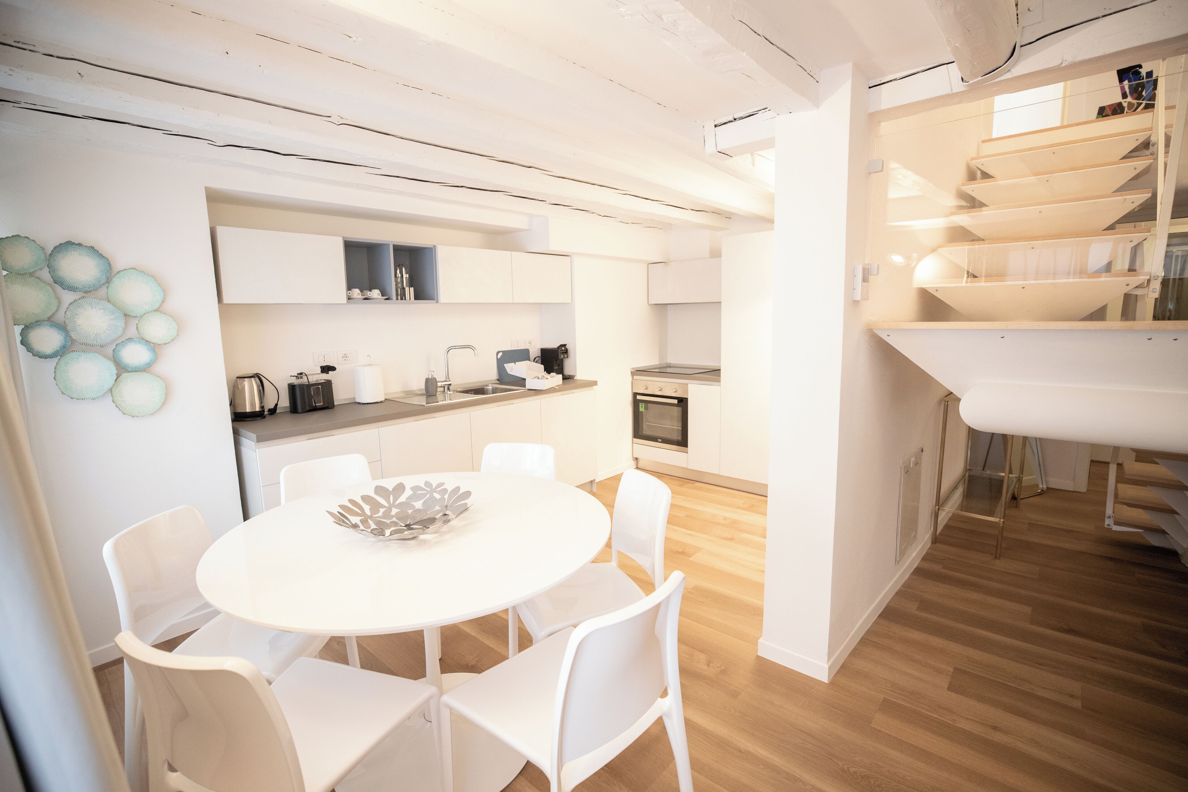 A modern kitchen with white cabinetry, a dining area with a white table and chairs, and a wooden staircase leading to an upper level.