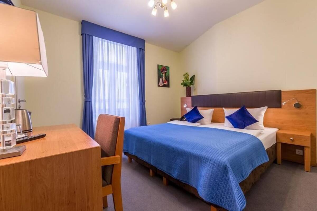 A hotel room with a bed, a desk with a lamp, a chair, a nightstand, and a window with curtains.