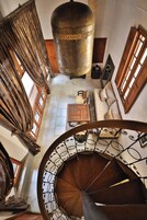Escadaria em espiral com corrimão trabalhado, um grande pilar de latão, um sofá e uma pequena mesa com um objeto decorativo.