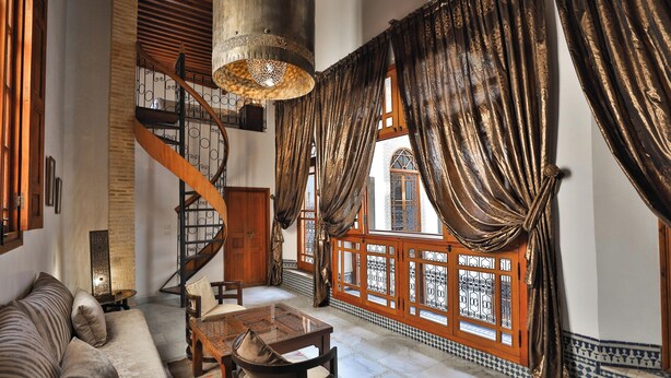 A spacious living area with a wooden staircase, a sofa, a dining table, and a large window with ornate curtains. - Riad Layla (Fes)