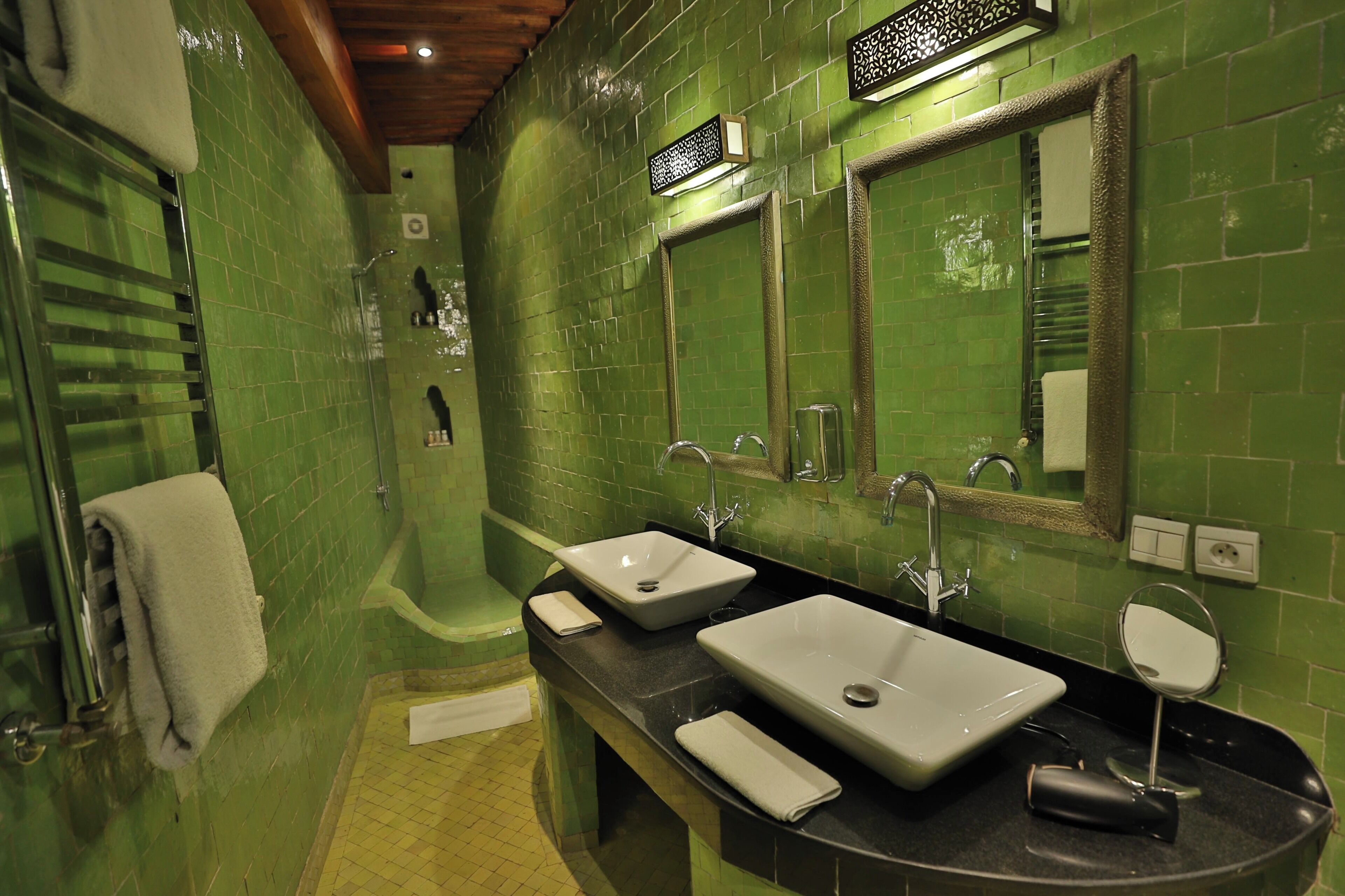 Un baño con paredes de azulejos verdes, dos lavamanos, una bañera y un espejo.