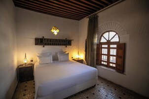 Quarto com uma cama grande, duas mesas de cabeceira com luminárias, uma prateleira decorativa na parede e uma janela com persianas de madeira.