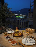 Una mesa dispuesta para una comida con varios platos, un tagine marroquí tradicional y vistas a un paisaje urbano al anochecer.