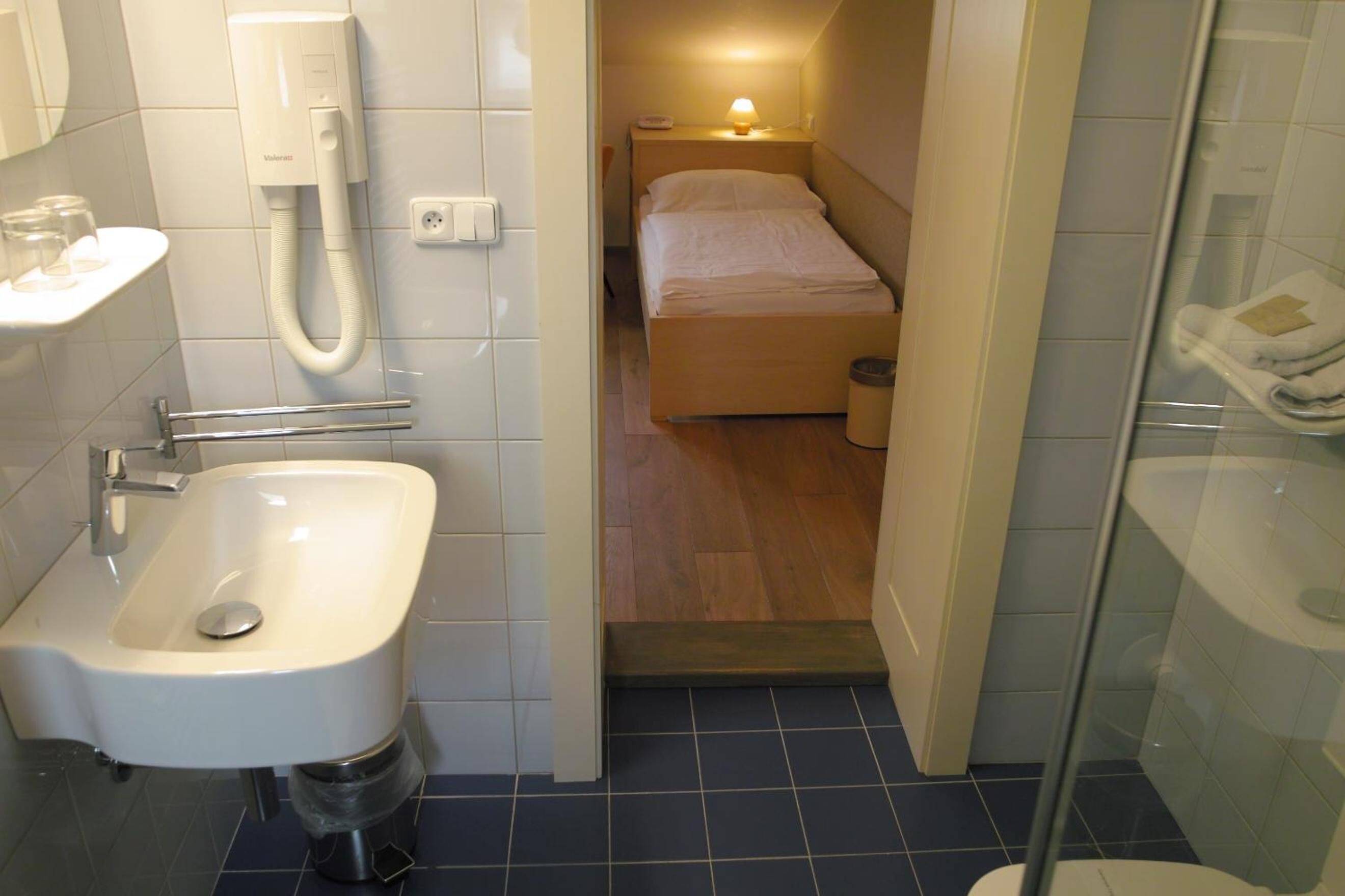 A compact hotel room with a bathroom, a bed, a bedside table with a lamp, and a telephone.