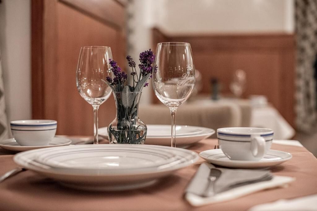 Una mesa de comedor dispuesta con platos, tazones, una taza y dos copas de vino, junto con un florero con flores.