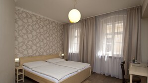 A double bed with a wooden frame, a bedside table with a lamp, a desk with a chair, and a window with sheer curtains. - Pension Krivá (Olomouc)