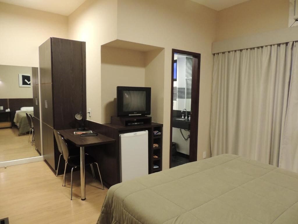 Una habitación de hotel con cama, escritorio con televisor, refrigerador pequeño y espejo.