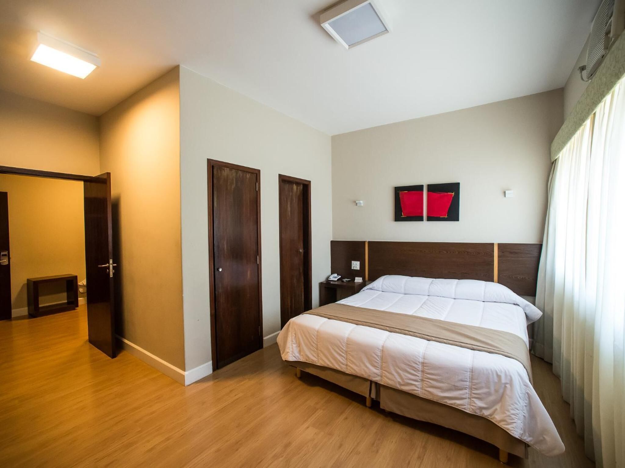 Quarto de hotel com piso de madeira, cama com roupa de cama branca, duas portas de madeira, mesa de cabeceira com abajur e luminária no teto.