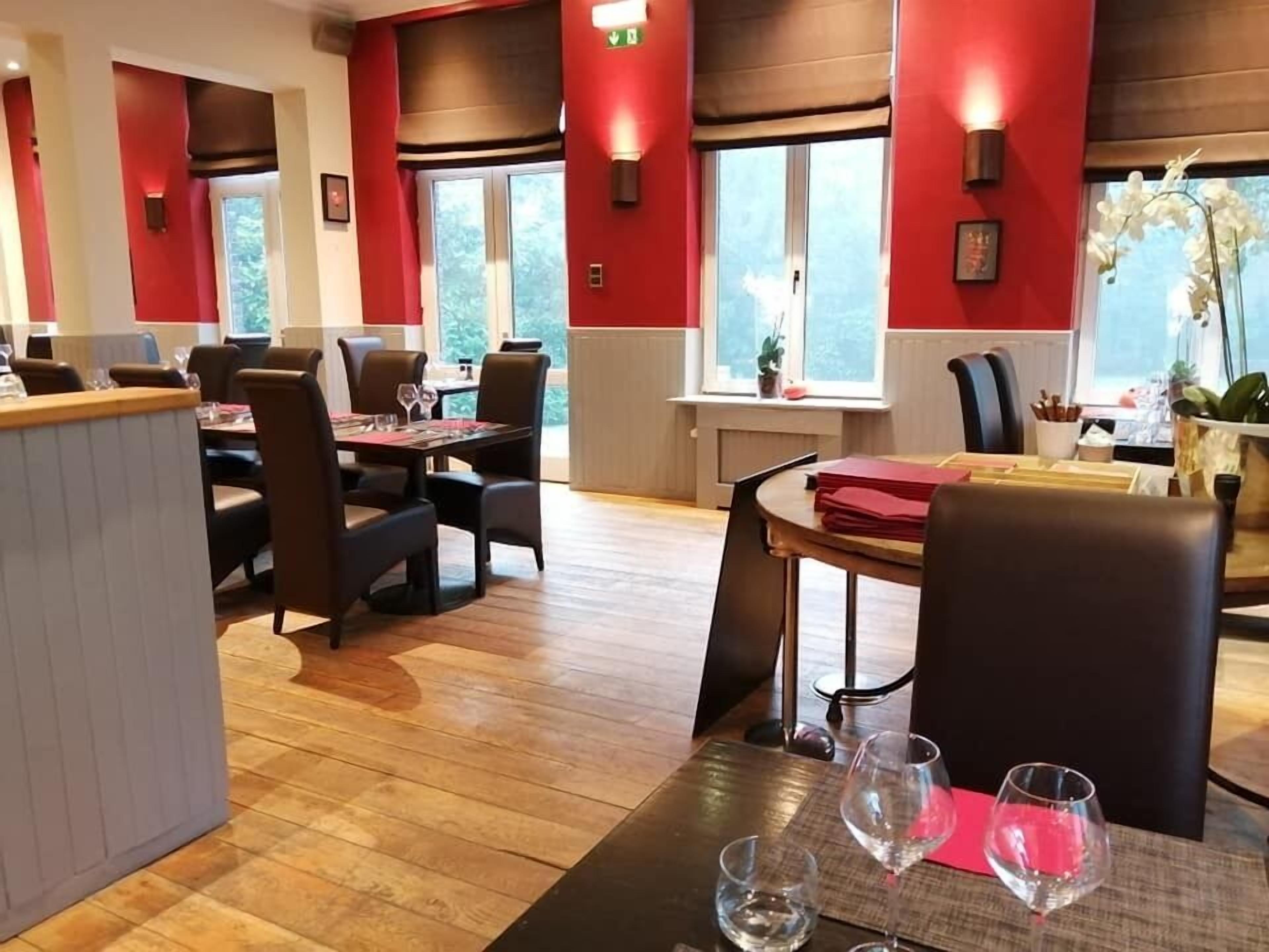 Un restaurant doté d’un plancher en bois, de murs rouges et de grandes fenêtres. Les tables sont disposées et accompagnées de chaises et de verres à vin.